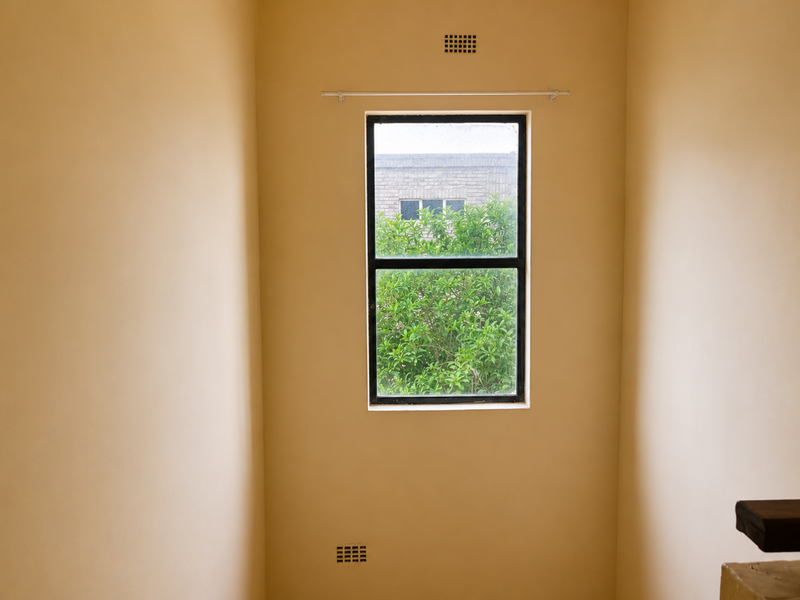 A simple, empty room featuring a metal-framed window with a curtain rod and wall-mounted ventilation grilles.