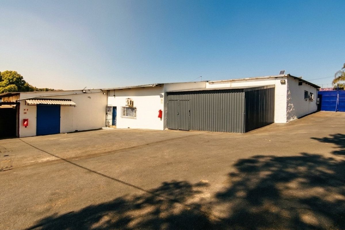 Rear exterior view of a mixed-use property featuring a large asphalt yard, corrugated metal roller doors, and functional security features.