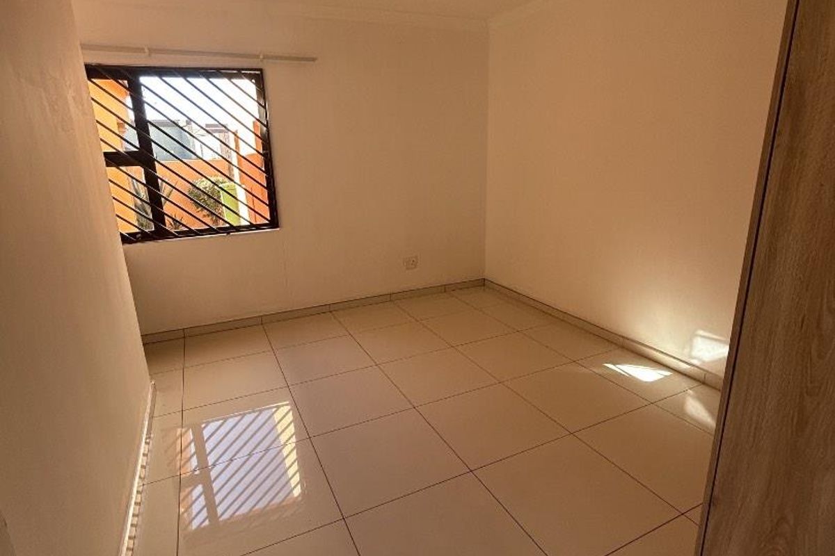 A clean, empty bedroom featuring neutral tiled flooring, a built-in cupboard, and a window fitted with security bars.