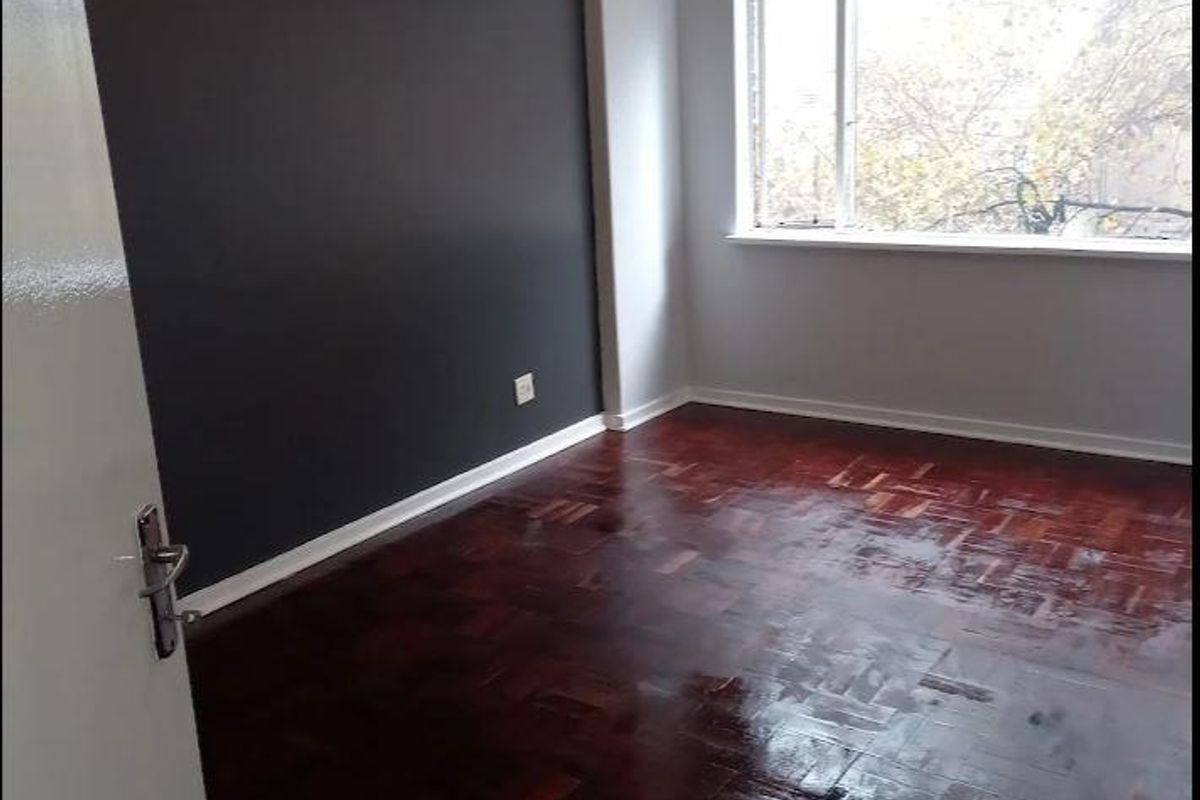 A bright, empty bedroom featuring polished parquet flooring, a large window providing natural light, and a dark accent wall.