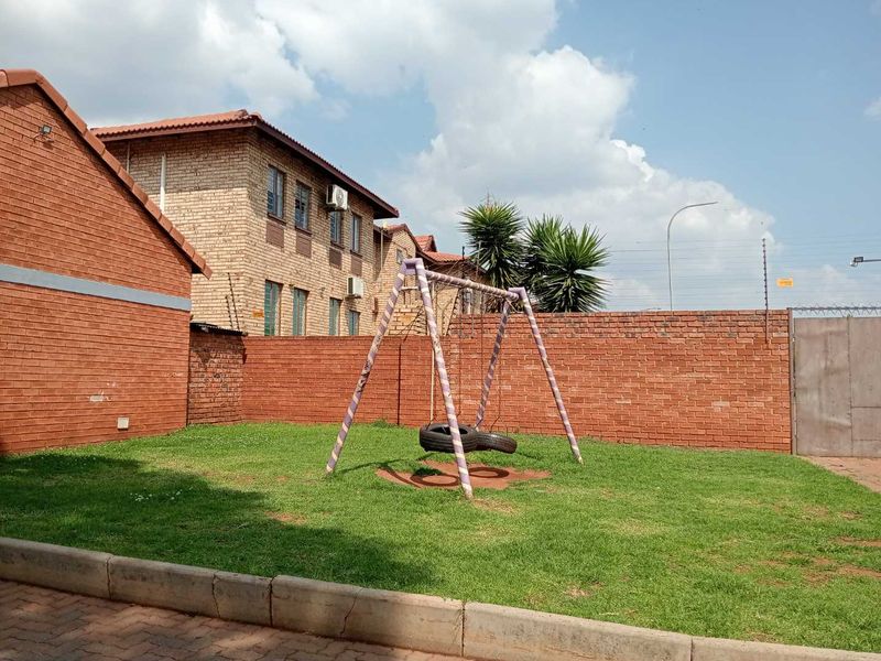 A communal outdoor area featuring a brick boundary wall with electric fencing, a grass lawn, and paved driveway access.