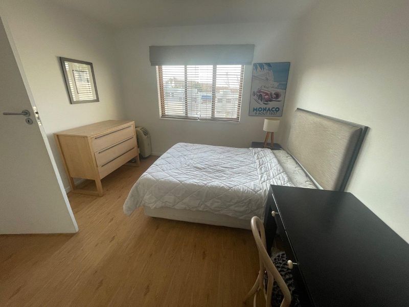 A well-lit bedroom featuring light-toned laminate flooring, neutral walls, and a window fitted with venetian blinds.