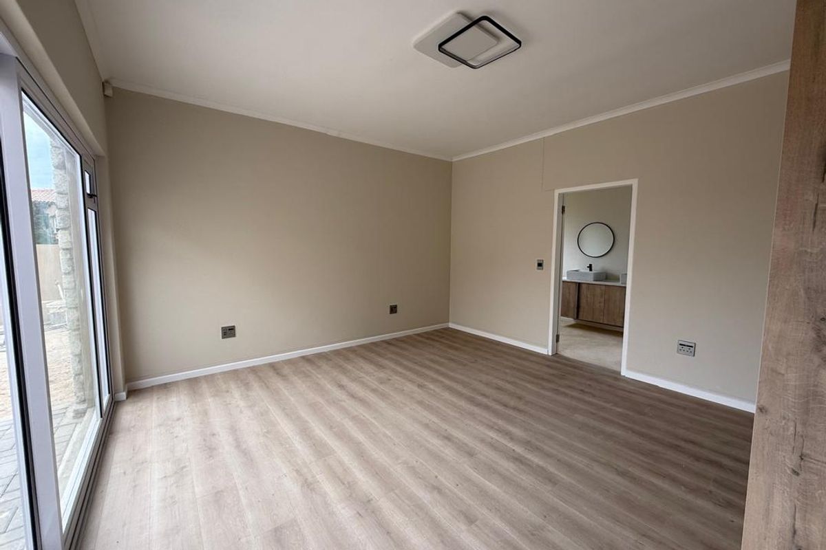A bright, empty bedroom featuring modern wood-look laminate flooring, neutral wall tones, and direct access to an en-suite bathroom.