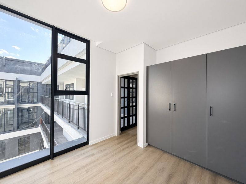 A bright, modern bedroom featuring large floor-to-ceiling windows, sleek grey built-in cupboards, and light laminate flooring, offering a clean and contemporary living space.