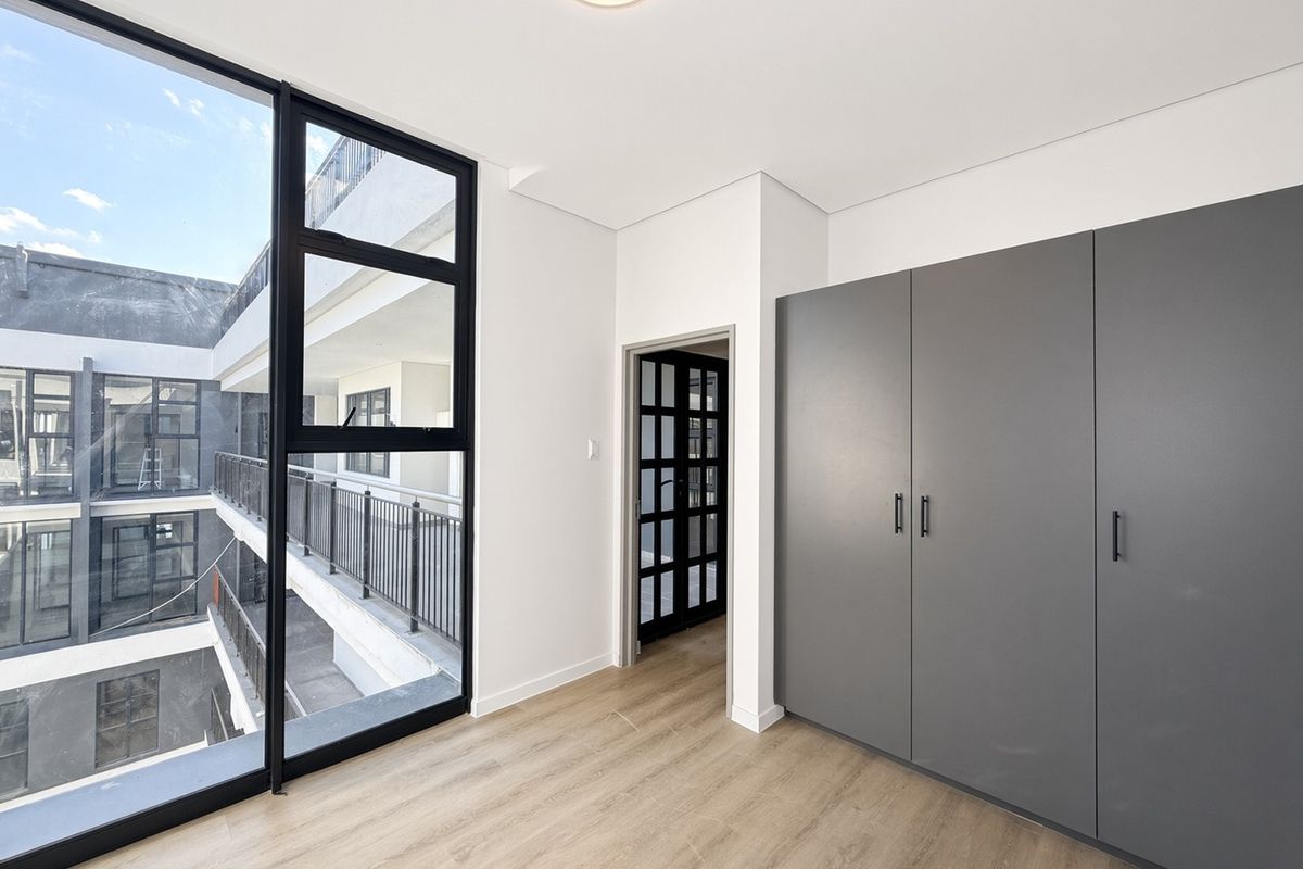 A bright, modern bedroom featuring large floor-to-ceiling windows, sleek grey built-in cupboards, and light laminate flooring, offering a clean and contemporary living space.