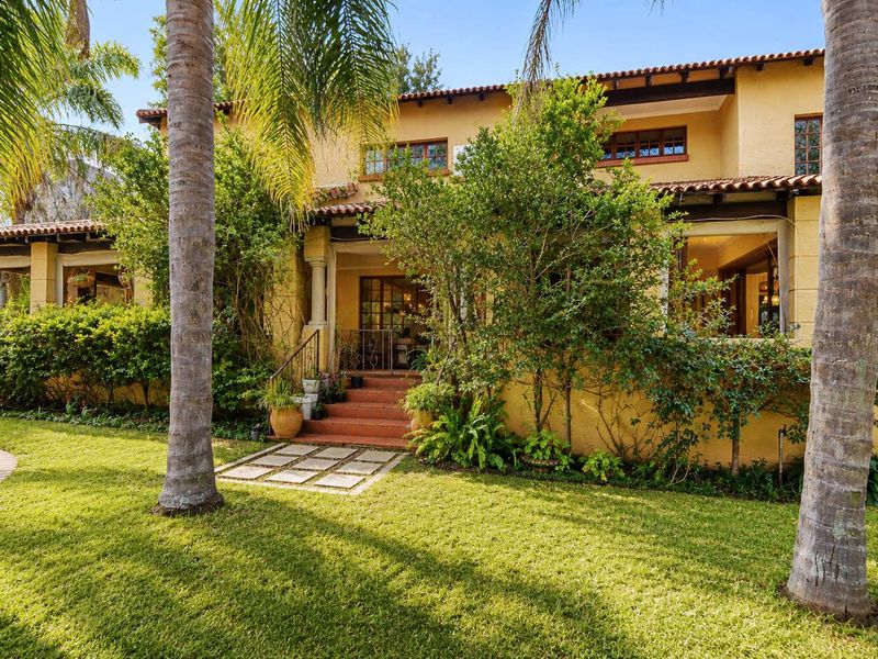 Rear view of a Mediterranean-style house featuring a covered patio, terracotta roof tiles, and a well-maintained garden with mature palm trees.