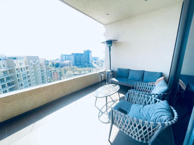 A spacious covered balcony featuring tiled flooring, a modern glass balustrade, and recessed ceiling lighting, offering an elevated urban view.