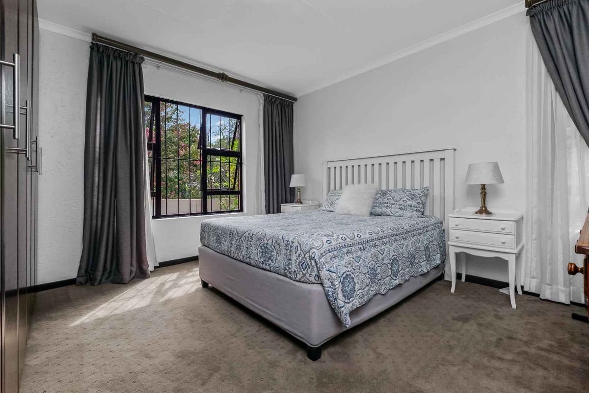A well-maintained bedroom featuring neutral carpeting, built-in cupboards, and a large window with security bars, offering a bright and comfortable living space.