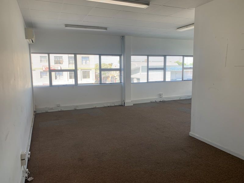 An empty, open-plan office space featuring large windows, carpeted flooring, suspended ceilings, and wall-mounted air conditioning.