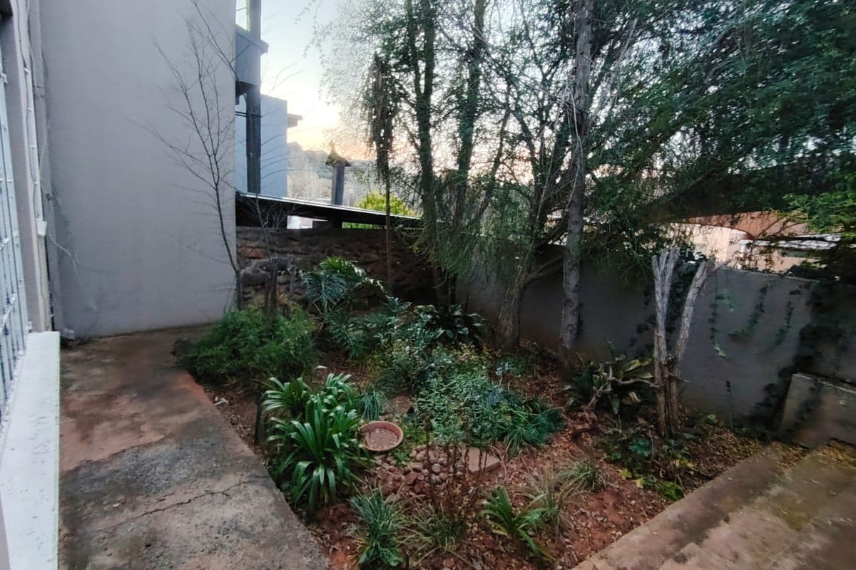 A rear garden area featuring established trees, landscaped planting beds, and concrete pathways adjacent to the property structure.