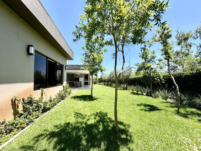 A well-maintained rear garden featuring a lush green lawn, mature trees, and a covered patio area attached to the main dwelling.
