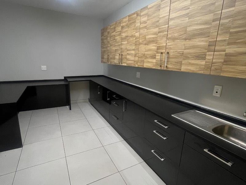 A clean, modern kitchen area featuring dark cabinetry, wood-grain upper cupboards, stainless steel sink, and neutral ceramic floor tiles.