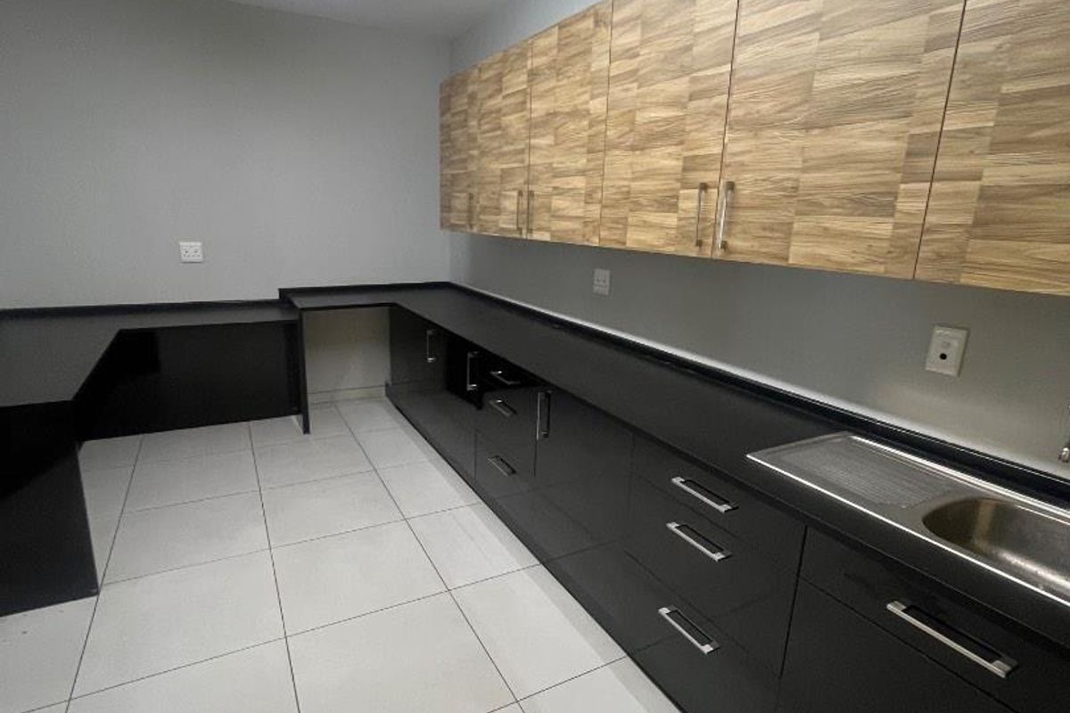 A clean, modern kitchen area featuring dark cabinetry, wood-grain upper cupboards, stainless steel sink, and neutral ceramic floor tiles.
