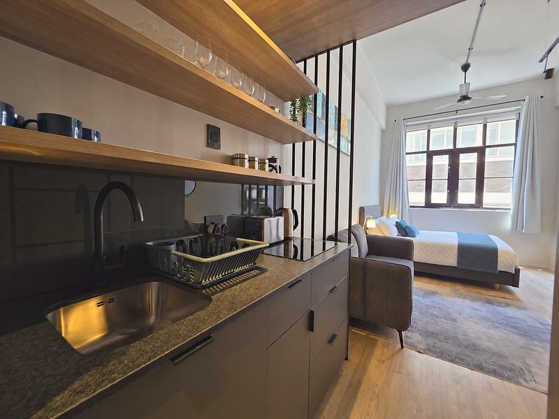 A modern open-plan studio apartment featuring a sleek kitchenette with stone countertops, induction hob, and timber shelving, seamlessly integrated with the bedroom area.