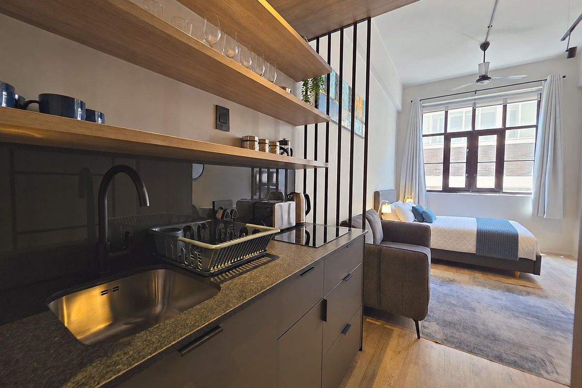 A modern open-plan studio apartment featuring a sleek kitchenette with stone countertops, induction hob, and timber shelving, seamlessly integrated with the bedroom area.