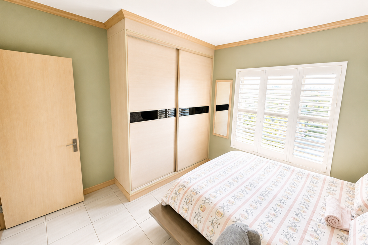 A well-maintained bedroom featuring built-in sliding door wardrobes, tiled flooring, and plantation shutters that allow for natural light.