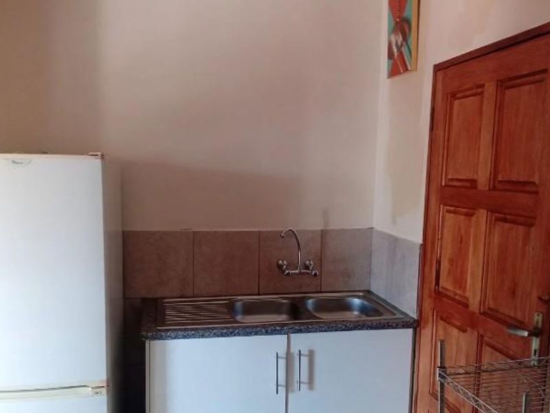 A compact kitchen area featuring a double bowl sink, tiled splashback, and functional cabinetry, with a wooden door leading to the adjacent space.