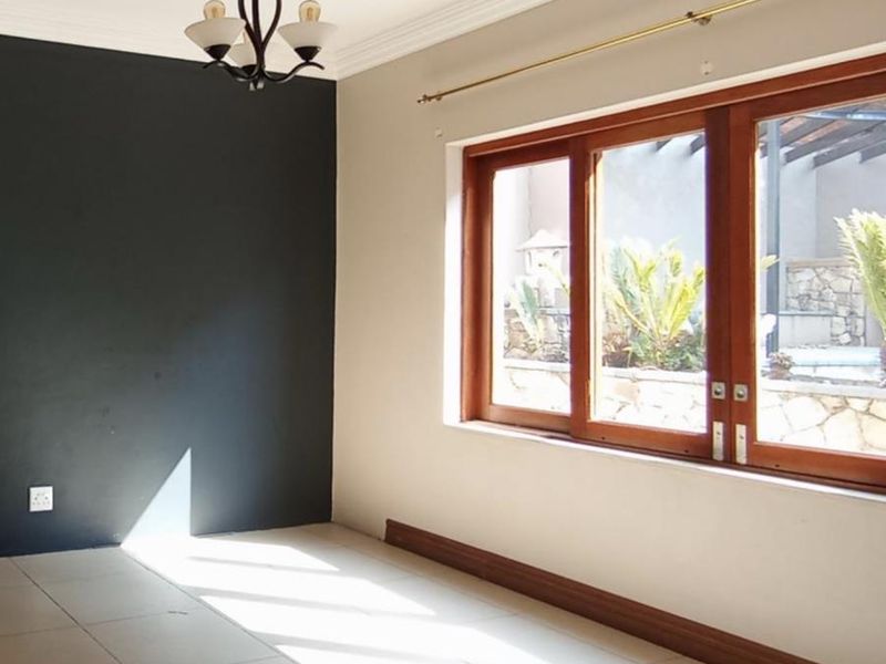 A bright, empty living area featuring tiled floors, a dark accent wall, and large wooden-framed windows overlooking the garden.
