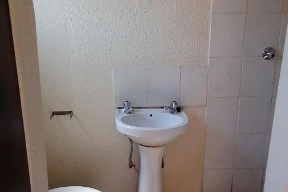 A compact bathroom featuring a pedestal basin, toilet, and a tiled shower area. The space requires cosmetic updates and floor refinishing.