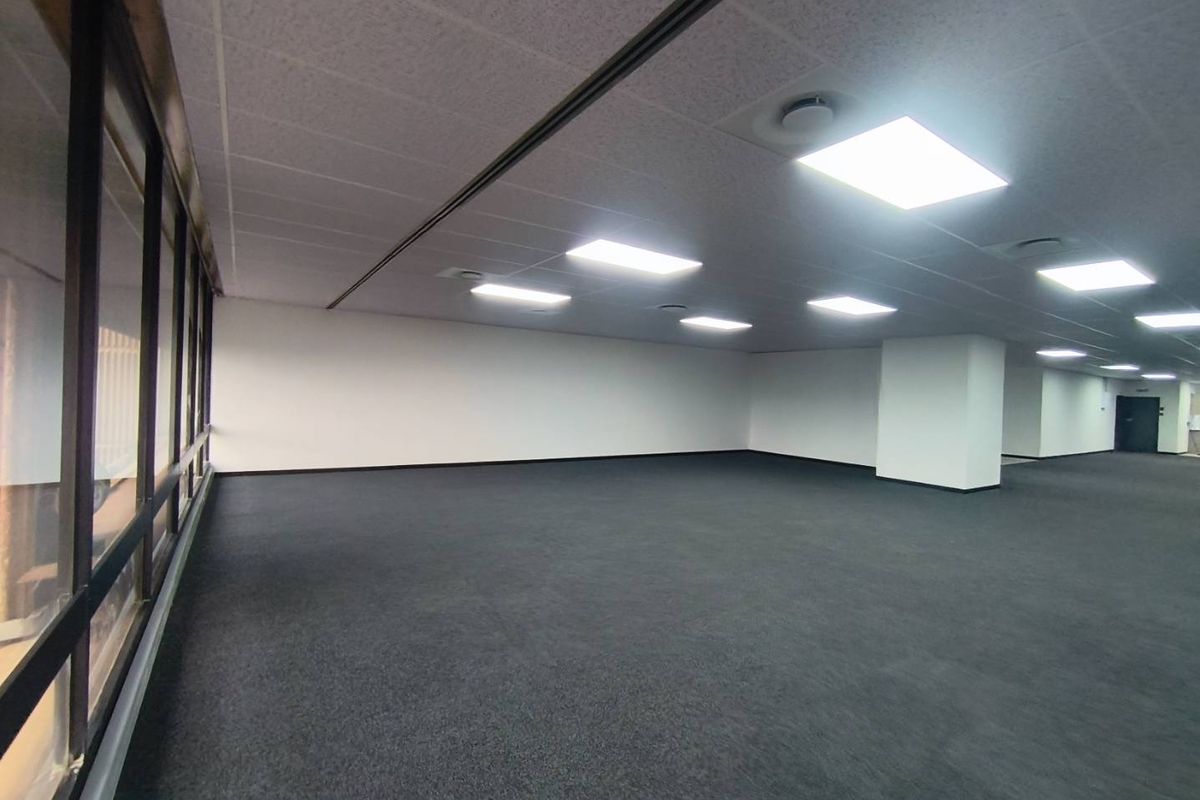 A spacious, empty open-plan office area featuring neutral carpeting, a suspended ceiling with recessed lighting, and large windows providing natural light.
