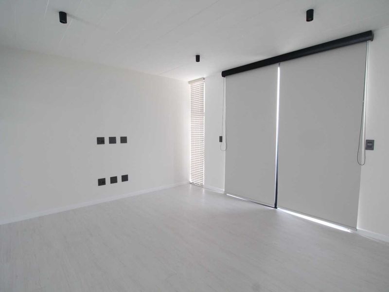 A bright, minimalist living room featuring neutral laminate flooring, recessed ceiling lighting, and modern roller blinds, offering a clean and versatile blank canvas.