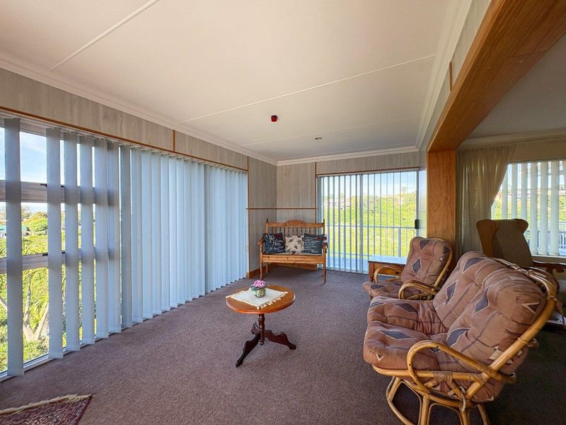 A sunlit living area featuring expansive windows with vertical blinds, carpeted flooring, and sliding glass doors leading to an outdoor space.