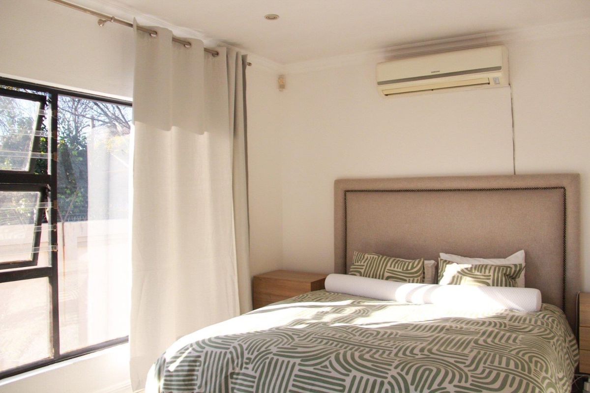 A well-lit bedroom featuring a wall-mounted air conditioning unit, large window with curtain rod, and recessed ceiling lighting.