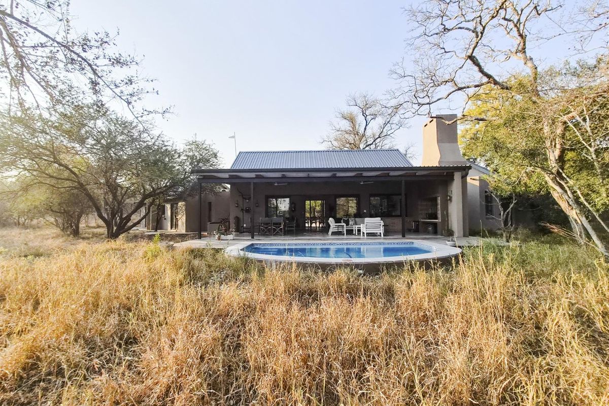 Rear view of the property featuring a covered patio, outdoor fireplace, and a swimming pool set within a natural bushveld landscape.