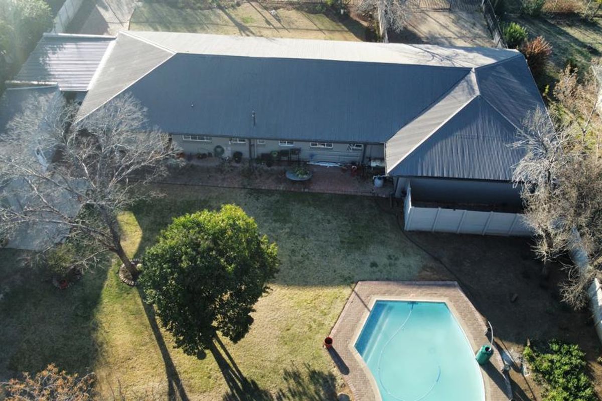 Aerial view of the property featuring a large metal roof, a spacious backyard with mature trees, and a paved swimming pool area.
