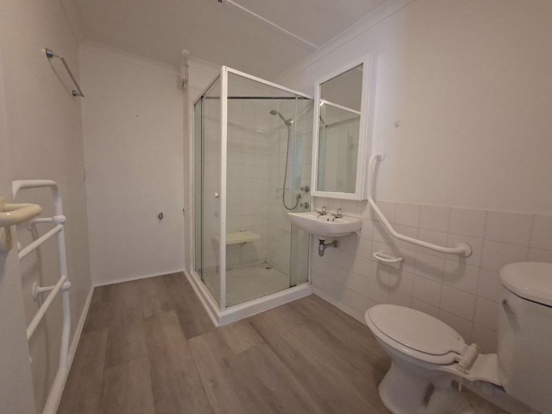 A functional bathroom featuring a shower enclosure, wall-mounted basin, toilet, and safety grab rails, finished with modern vinyl flooring.