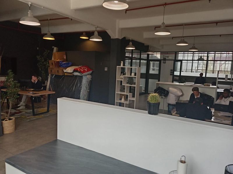 Open-plan office space featuring industrial-style exposed ceilings, pendant lighting, and large windows providing ample natural light.
