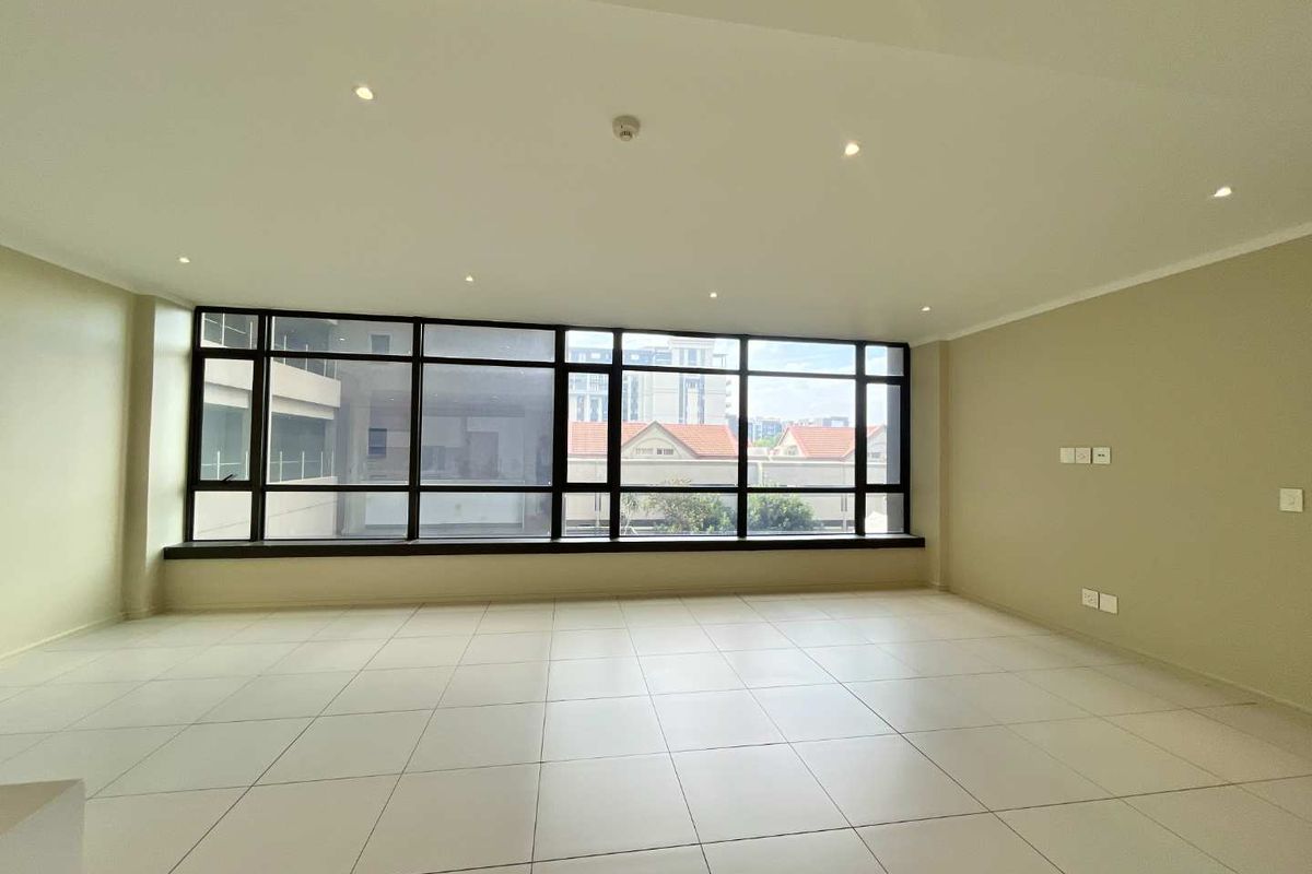 A bright, spacious, and empty living area featuring large floor-to-ceiling windows, neutral tiled flooring, and recessed ceiling downlights.