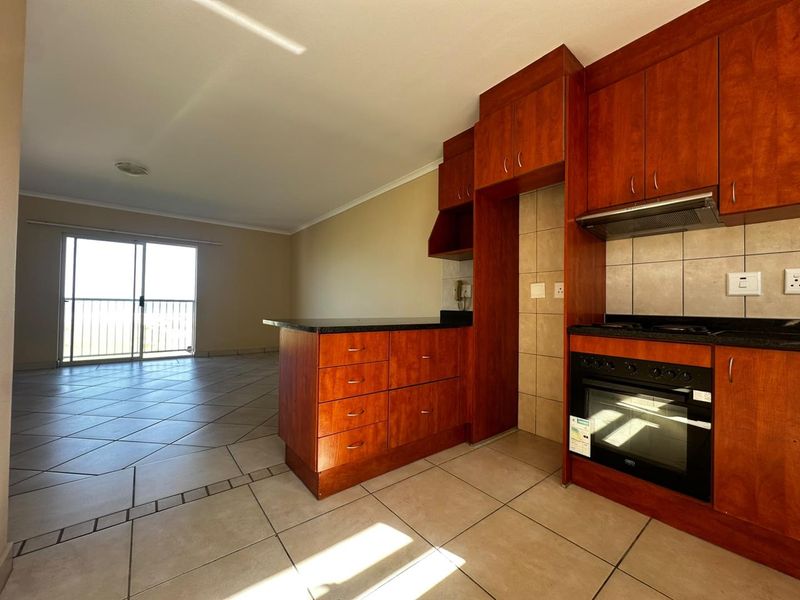 Open-plan kitchen and living area featuring warm-toned wooden cabinetry, granite countertops, built-in oven, and direct access to a balcony through sliding glass doors.