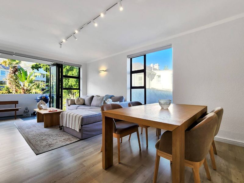 Bright open-plan living and dining area featuring modern laminate flooring, track lighting, and large black-framed windows leading to a private balcony.