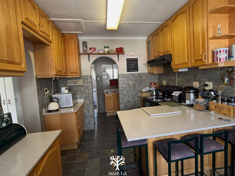 A functional kitchen featuring wooden cabinetry, stone countertops, tiled backsplash, and stone flooring, with an open archway leading to an adjacent utility area.