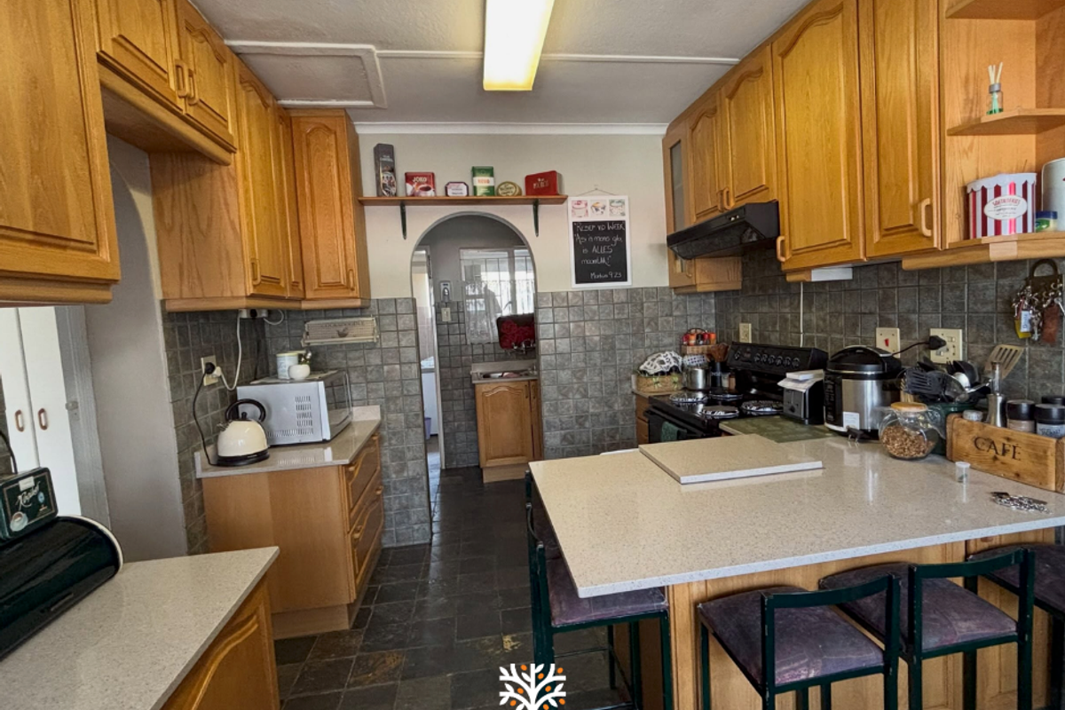 A functional kitchen featuring wooden cabinetry, stone countertops, tiled backsplash, and stone flooring, with an open archway leading to an adjacent utility area.