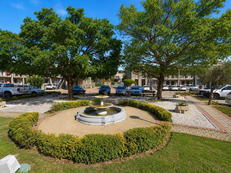 A well-maintained office park courtyard featuring a central fountain, mature trees, landscaped garden beds, and paved parking areas.