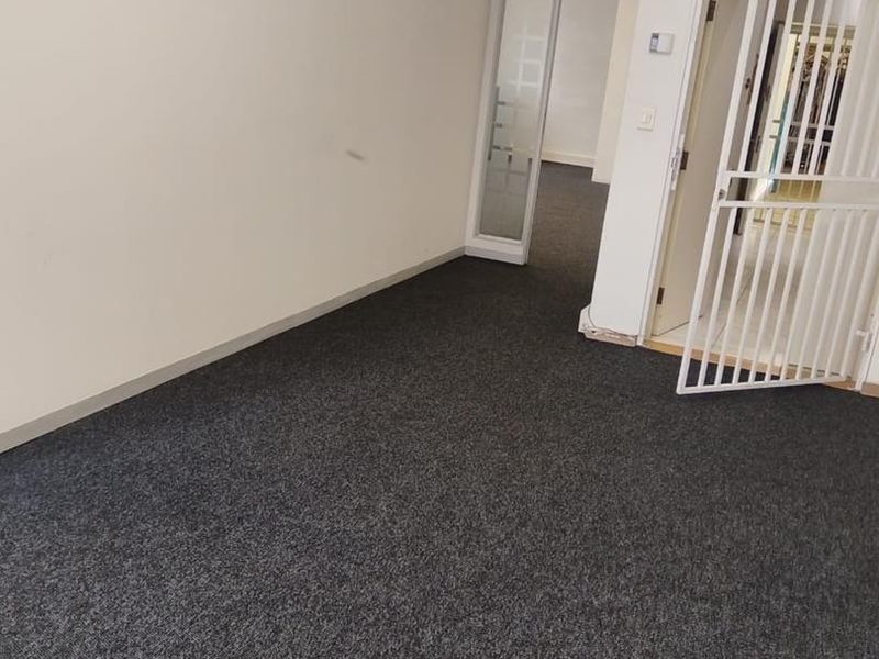 An empty office space featuring dark carpeting, exposed ceiling ducting, a glass partition, and a white security gate at the entrance.