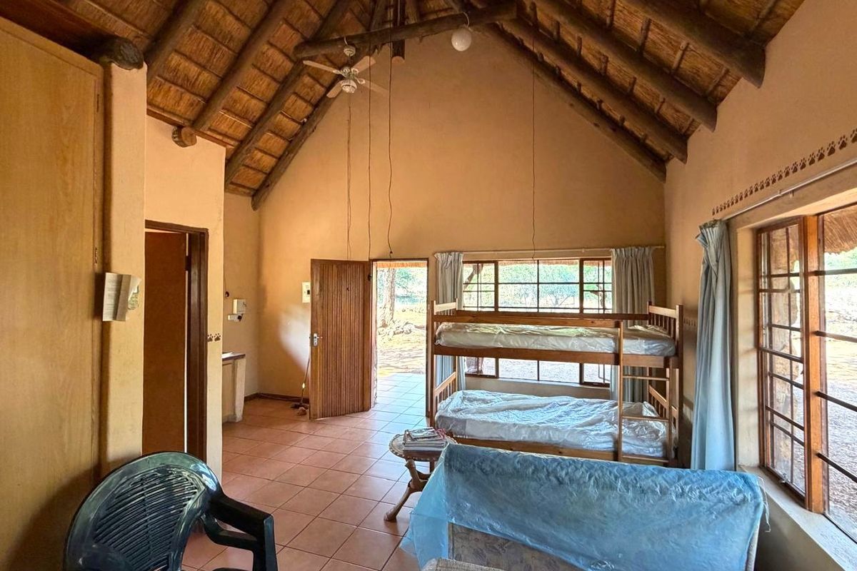 A rustic bedroom featuring a high vaulted thatched ceiling with exposed timber rafters, tiled flooring, and large windows providing natural light.