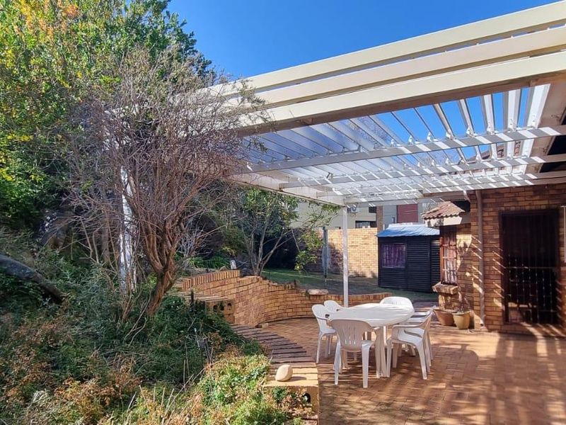 A covered brick-paved patio area featuring a louvred roof structure, adjacent to a brick exterior wall and landscaped garden area.