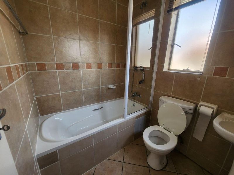 A functional bathroom featuring a bathtub with a shower screen, a toilet, and a wall-mounted basin, finished with neutral-toned wall and floor tiles.