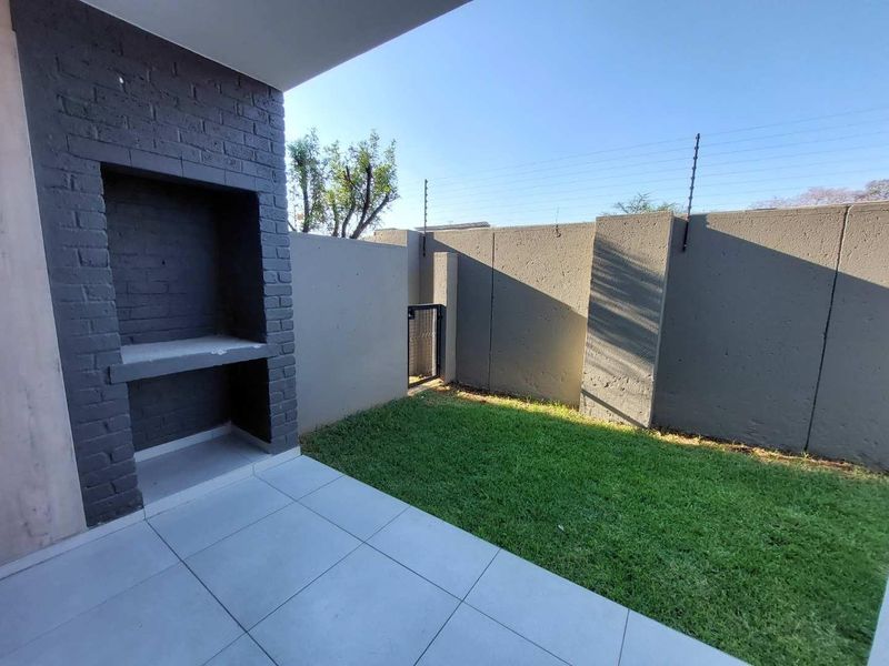 A private outdoor patio featuring a built-in braai, tiled flooring, and a small, well-maintained lawn enclosed by a secure perimeter wall with electric fencing.