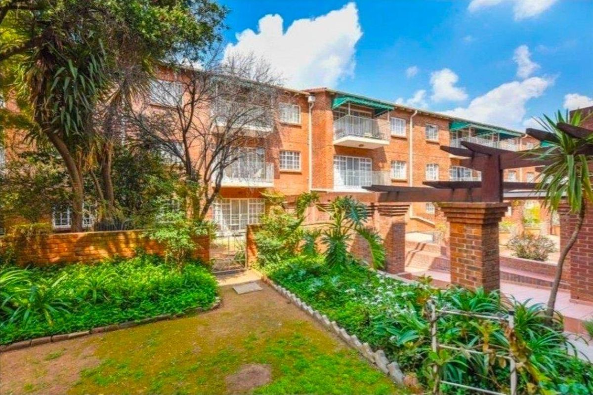 Rear view of a multi-story brick apartment complex featuring private balconies, a landscaped garden area, and a decorative brick pergola.