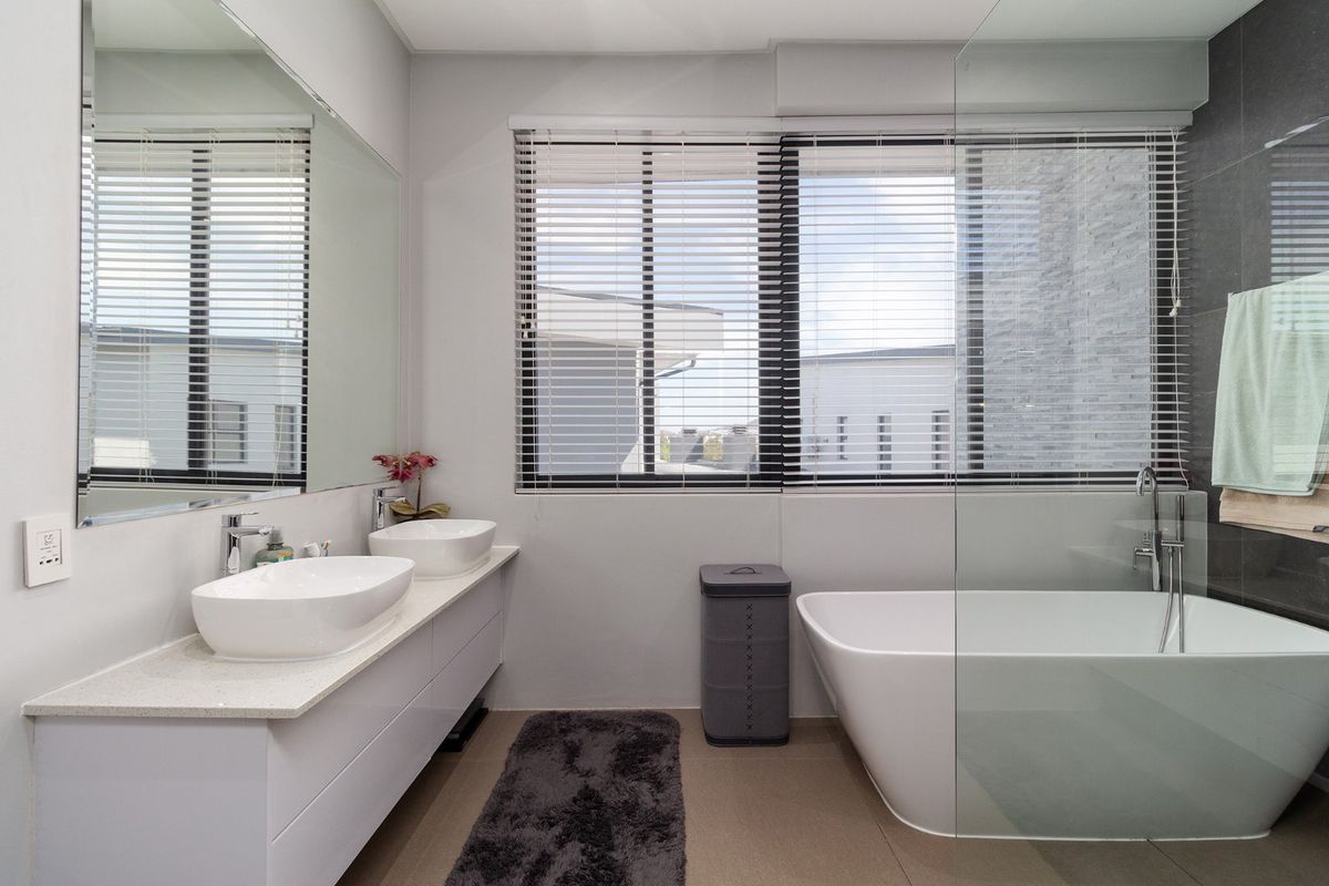 Modern bathroom featuring a double vanity with vessel sinks, a sleek freestanding bathtub, a glass shower screen, and large windows with venetian blinds.