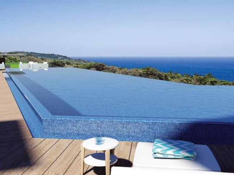 An infinity edge swimming pool featuring blue mosaic tiling and timber decking, offering expansive ocean views.