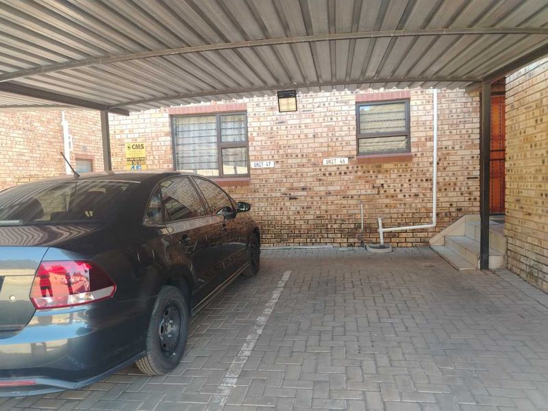 A covered carport area with brick facade, paved flooring, and external security lighting, providing dedicated parking space for the apartment unit.