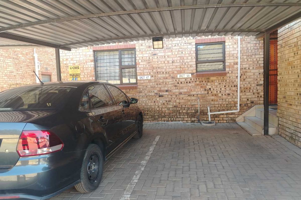 A covered carport area with brick facade, paved flooring, and external security lighting, providing dedicated parking space for the apartment unit.