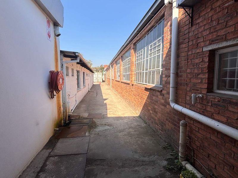 A narrow exterior service alleyway between two industrial buildings, featuring exposed brick walls, steel-framed windows, and a concrete walkway.