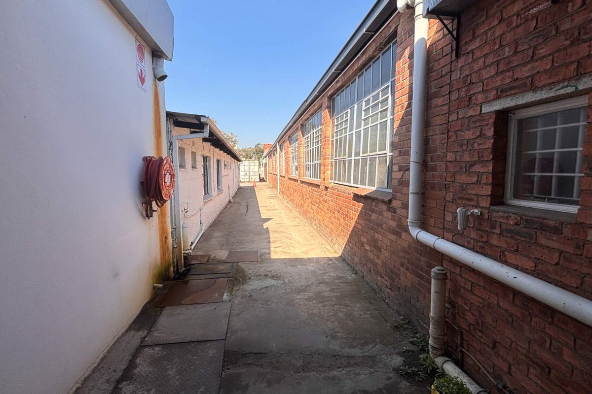 A narrow exterior service alleyway between two industrial buildings, featuring exposed brick walls, steel-framed windows, and a concrete walkway.