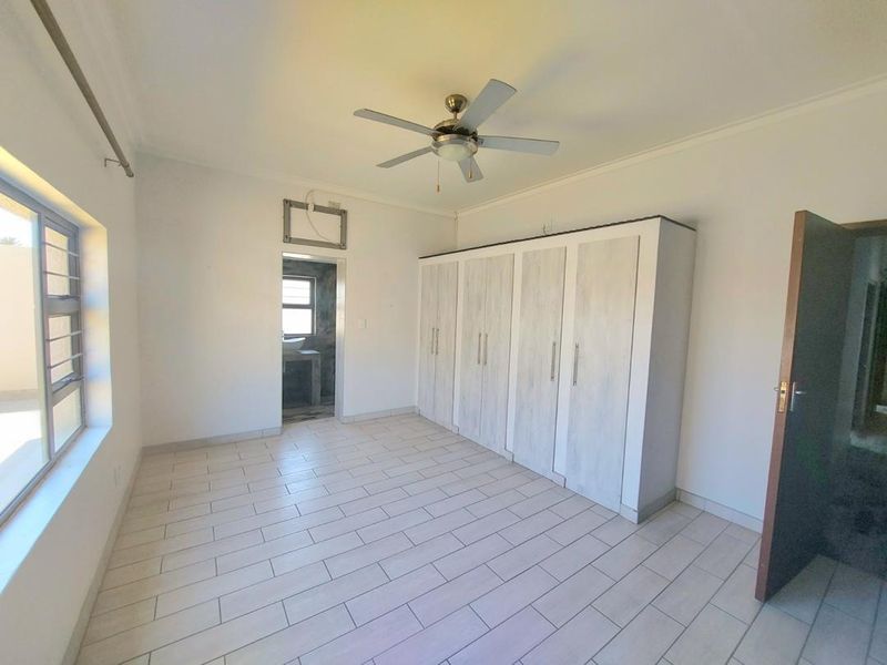 A bright, empty bedroom featuring neutral tiled flooring, built-in cupboards, a ceiling fan, and an en-suite bathroom entrance.